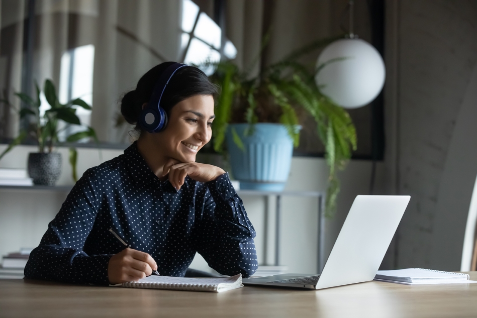 A person sitting and smiling in front of a laptop, wearing headphones and holding a pen to a notepad.